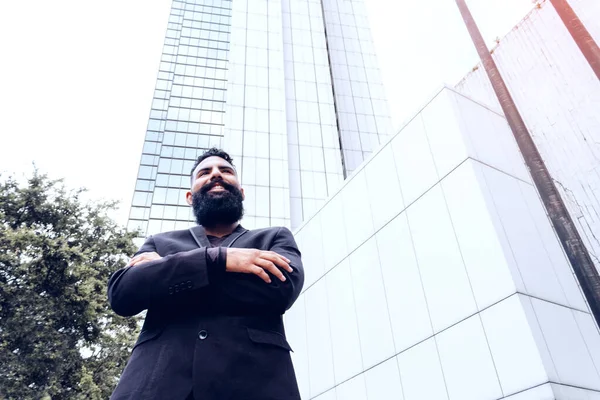 business executive man businessman in black suit caucasian with long beard crossing arms in modern building structure outdoor smiling in city.