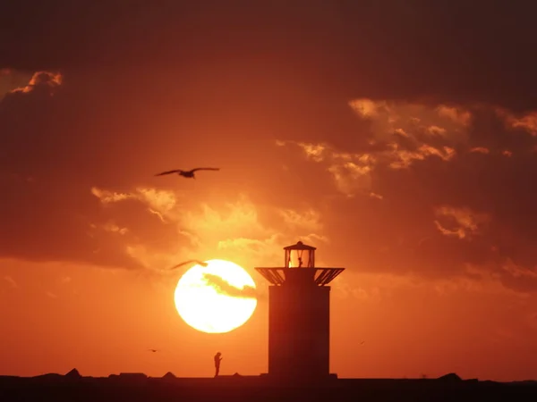 Bir deniz fenerinin yanında gün batımını izleyen bir insan silueti.