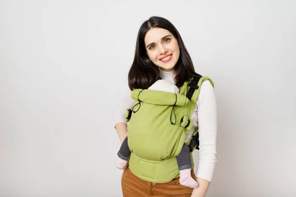 Bebek taşıyıcısında beyaz bir arka planda bebeği olan neşeli genç bir anne.