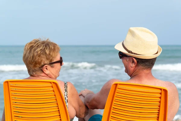 当一对年长的夫妇坐在海滩上时 他们彼此凝视着对方并牵着手的背影 放宽暑假的限制 — 图库照片