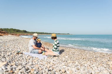 Baba, anne ve küçük kızları Akdeniz kıyısındaki çakıl taşı plajında oturuyorlar. Rahatlatıcı ve huzurlu tatiller.