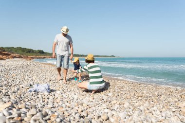 Küçük çocuk babasının elini tutuyor ve annesine doğru yürüyor. Akdeniz sahilinde kollarını açmış onu bekliyor. Yaz titreşimleri.
