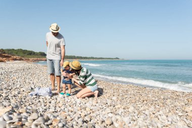 Annesi küçük kızına sarılırken babası çakıl taşı plajında büyük bir gülümsemeyle onlara bakıyor. Yaz tatilleri. 