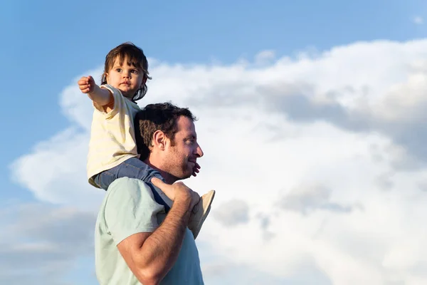 Little girl sitting on her father's shoulders with open arm and looking at the camera direction on a background of blue sky with clouds. Bonding and love concept.