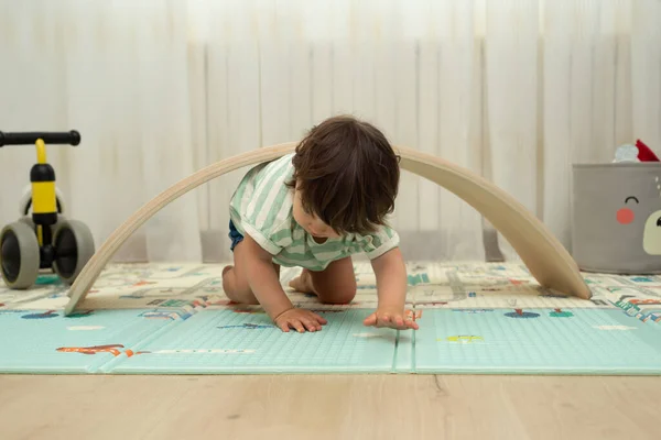 Toddler crawling under balance board in a playroom.