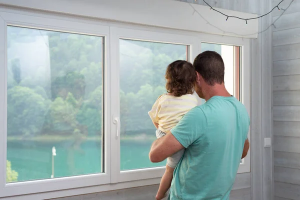 Rear view of a father holding his little girl on arms and enjoying a lake views from the window of his apartment.