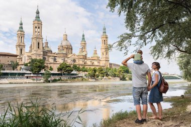 Back view of a young family, parents and baby, doing sightseeing in Zaragoza. They are standing at Ebro river bank and enjoying Basilica del Pilar view. Family holidays.
