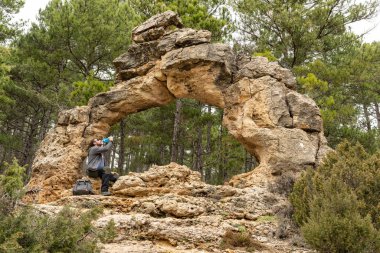 Young man is hydrating during a trekking while is sitting on a natural stone arch located in the middle of a pine tree forest. Active lifestyle. 
