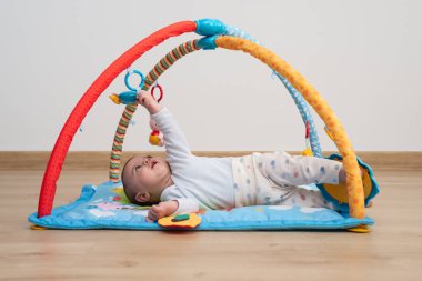 5 months baby lying face up on a blue play mat while trying to catch a toy from a baby gym. Montessori activities.