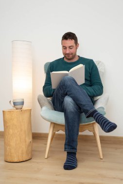 Young man reading a book in a comfortable reading corner. Hygge home interior.
