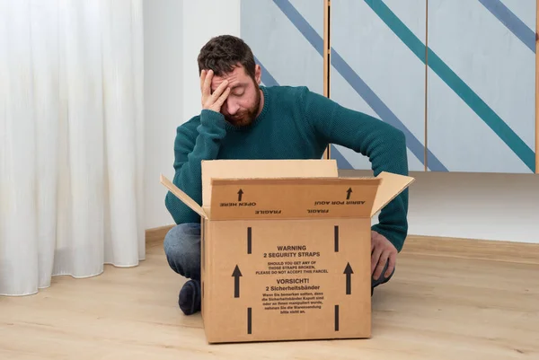 Sad young man sitting on wooden floor at home, looking inside an open package and feeling desperate about received a wrong item.