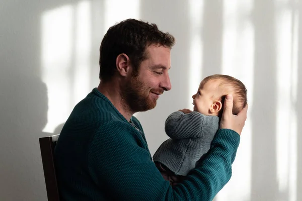 Young father and his baby looking at each other while smiling, sitting in sunlight with blinds shadows. Bonding and fatherhood concept.