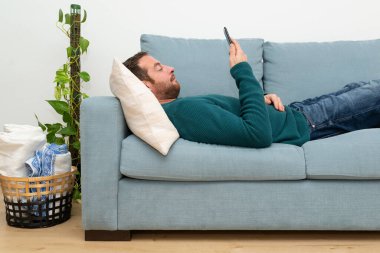 Young guy lying on a blue sofa and using his smartphone. 