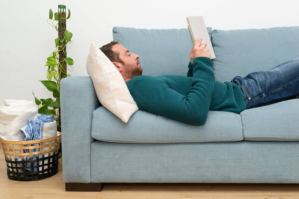 Side view of a attractive, bearded man lying on a sofa and reading a novel. Relaxing time. 