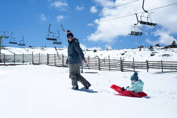 Smiling father on ski wear walking and pulling a red sledge with her little girl sitting on it at a ski resort.