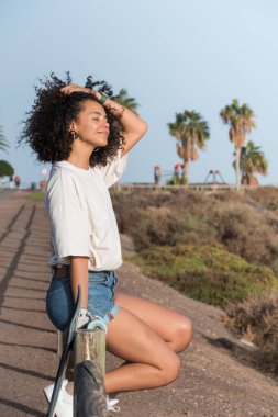 Afro saçlı genç patenci kadın çitin üzerinde oturuyor esintiyi ve güneşi yüzünde hissediyor