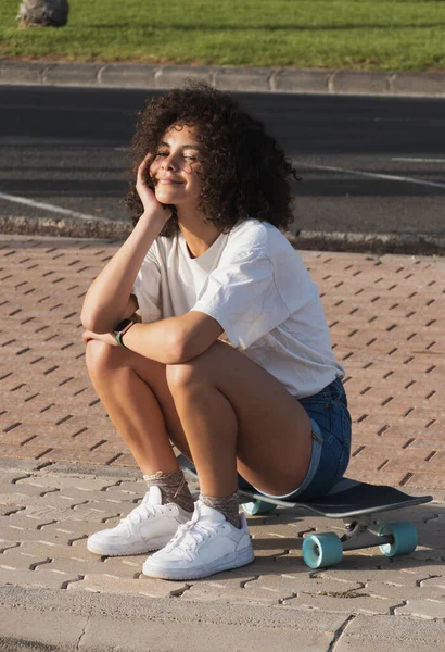 Açık havada kaykayında oturan afro saçlı genç bir kadın.