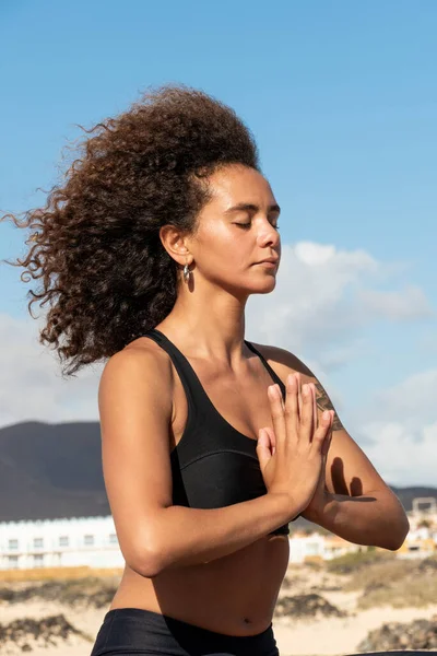 Açık havada yoga pozisyonunda oturan Afro genç kadın portresi. Elleri birarada ve gözleri kapalı.