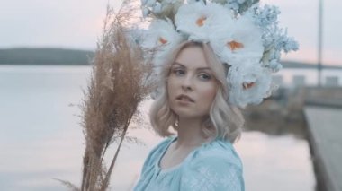 Kafasında çelenk olan Ukrayna vatandaşı bir kadın. Nakış. Nakış. Nehir manzarası. Elinde sarı çimen olan bir kadın.