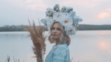 Kafasında çelenk olan Ukrayna vatandaşı bir kadın. Nakış. Nakış. Nehir manzarası. Elinde sarı çimen olan bir kadın.