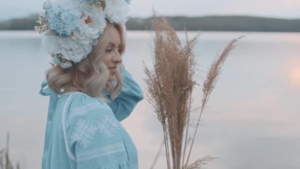 Женщина в украинском национальном костюме с венком на голове. Вышивка. Вид на реку. Женщина с жёлтой травой в руках. Медленное движение — стоковое видео