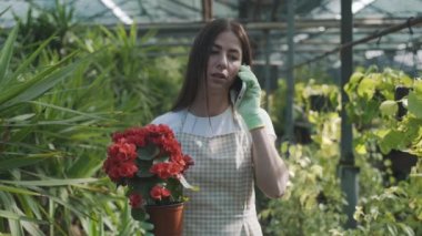 Serada elinde çiçeklerle genç bir kız telefonda bitkilerin arka planına karşı konuşuyor. Yavaş çekim
