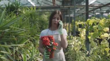 Serada elinde çiçeklerle genç bir kız telefonda bitkilerin arka planına karşı konuşuyor. Yavaş çekim