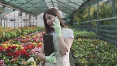 Elinde dosyasıyla serada elinde notlar için bekleyen genç bir kız telefonda bitkilerin arka planında konuşuyor. Yavaş çekim
