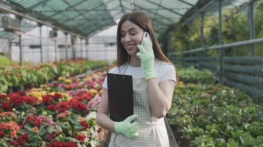 Elinde dosyasıyla serada elinde notlar için bekleyen genç bir kız telefonda bitkilerin arka planında konuşuyor. Yavaş çekim