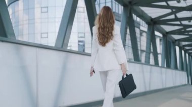 Young confident business woman in white suit walking with bag of laptop. Busy beautiful girl leaving office building after work. Slow motion