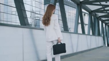Young confident business woman in white suit walking with bag of laptop. Busy beautiful girl leaving office building after work