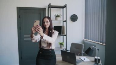 Genç sekreter, iş kadını, gömlekli ve etekli, bilgisayarlı masaya yaslanmış cep telefonuyla selfie çekimi yapıyor. İş içeriği