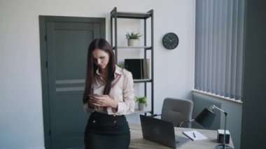 Genç sekreter iş kadını gömlekli ve etekli. Ofiste cep telefonundan bir şey arıyor. Farklı duygular gösteriyor. İş içeriği