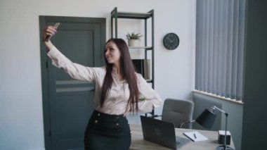 Genç sekreter, iş kadını, gömlekli ve etekli, bilgisayarlı masaya yaslanmış cep telefonuyla selfie çekimi yapıyor. İş içeriği
