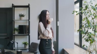 Genç iş kadını ofiste duruyor ve kahve içiyor. İş içeriği