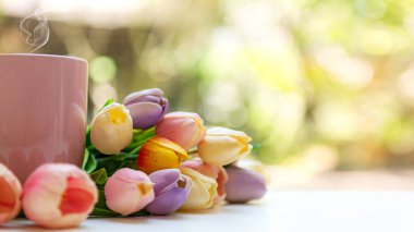 Lale çiçekleri ve yeşil arka planı bulanık olan beyaz ahşap pencereli pembe buharlı kahve fincanı.