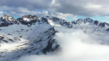 Dik kayalıklardan oluşan karlı, bulutlu dağlar. Ulaşılması zor zirveler.