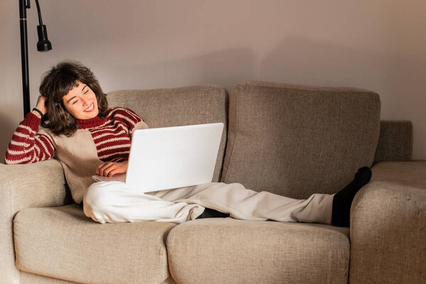 A young brunette woman is watching a movie on her computer lying on her sofa in her living room at home