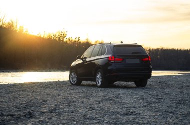 Büyük siyah ve mavi bir SUV sınıfı araba günbatımında ışıkları açık sarı güneş ışığıyla kamufle edilmiş bir nehir kıyısında duruyor.