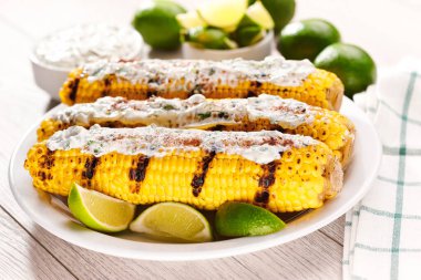 Elotes, grilled corn, roasted cobs are covered in creamy mayonnaise, seasoned with chili powder and cilantro, and  lime on a white table.