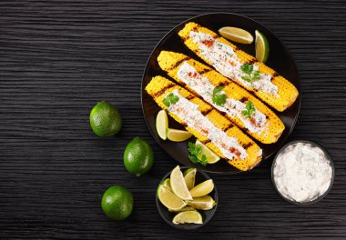 Flat-lay concept of grilled sweet corn with lime and sauce over dark background, top view. Vegetarian, healthy, clean eating. Summer vegan dinner or snack.