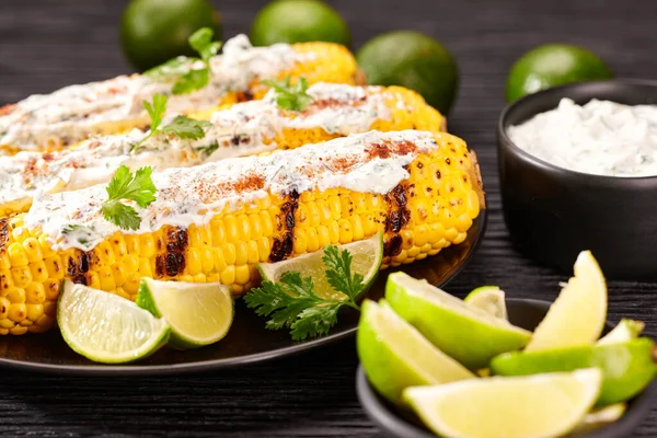 Grilled corn on the cob with sauce, coriander, and lime on a dark table. Mexican food. Corn grilled with sauce.