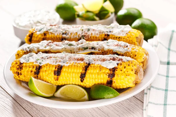 Elotes, grilled corn, roasted cobs are covered in creamy mayonnaise, seasoned with chili powder and cilantro, and  lime on a white table.