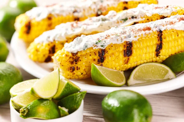 Grilled corn on the cob with sauce, coriander, and lime on a light table, selective focus. Elote on a white plate, grilled mexican street corn. Vegetarian, healthy, clean eating.