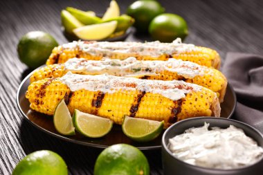 Roasted or grilled sweet corn cobs with with cream sauce, lime and cilantro, selective focus. Mexican street corn on dark table.