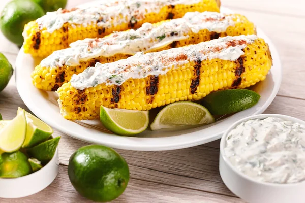 Grilled corn on the cob with sauce, coriander, and lime on a white table, selective focus. Elote on a white plate, grilled mexican street corn.
