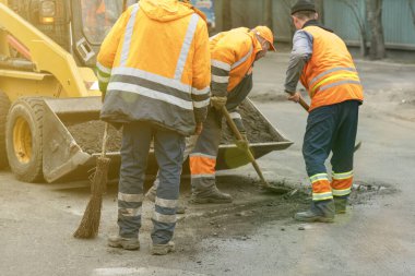 Parlak turuncu yansıtıcı üniformalı yol işçileri birikmiş kumu kazmak için kürek kullanırlar. Yol ve otoban kaldırımlarının bakımı. Çukur onarımı işe yarıyor. ses tonlu