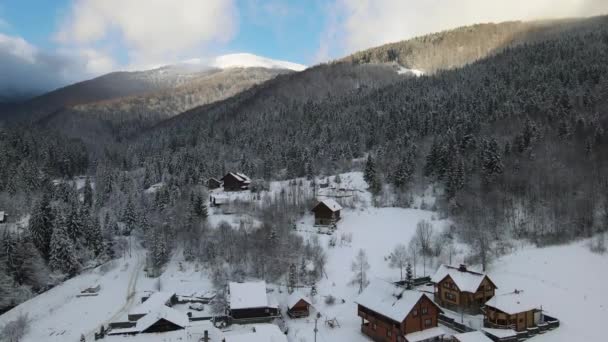 Εναέρια χειμερινό τοπίο με μικρά αγροτικά σπίτια μεταξύ χιονισμένο δάσος σε κρύα βουνά. — Αρχείο Βίντεο