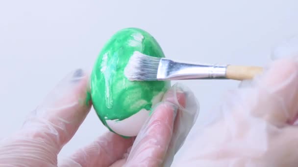 Närbild av en person som målar påskägg med en speciell pensel. En flicka målar ett påskägg grönt och vitt. Förberedelser inför påsk. Vitt bord, närbild. — Stockvideo