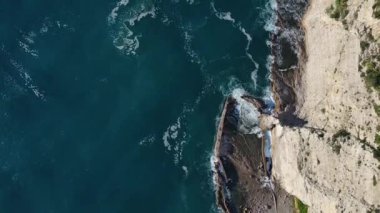 Dalgaların yukarıdan görünüşü mavi bir okyanusta kayalara çarpıyor. Deniz dalgaları güzel sahil manzaralı insansız hava aracı. Kuşların bakış açısı okyanus dalgalarının yukarıdan kayalıklara çarpmasını gösteriyor..